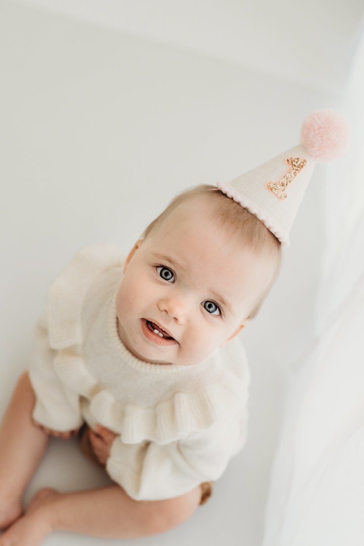 A young child with blue eyes and light hair wearing a white sweater and a pink party hat with a gold number 1, sitting on a light surface against a white background.