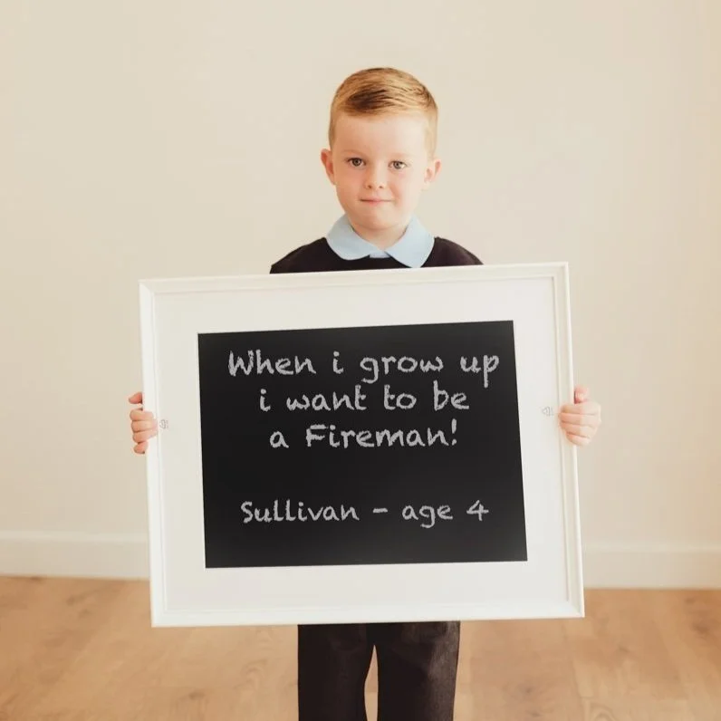 A young boy standing indoors, holding a white-framed blackboard with a handwritten message about wanting to be a firefighter when he grows up, and his name and age.