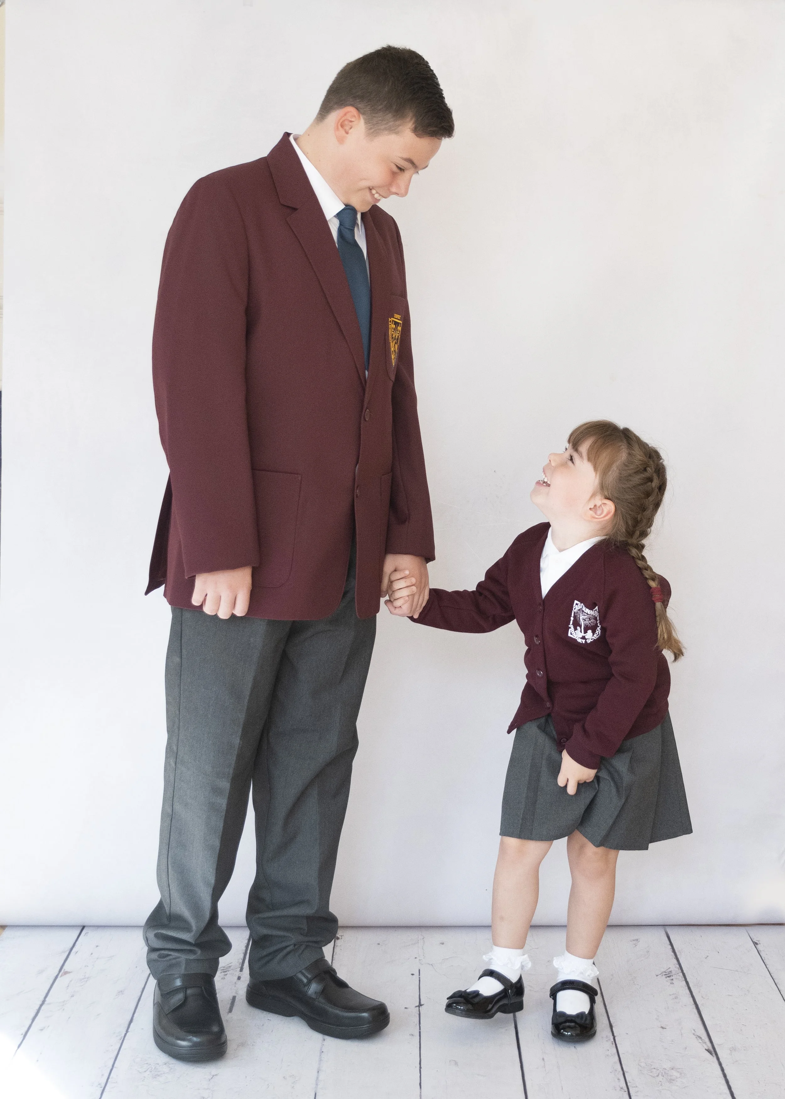 A tall young man in a school blazer smiling and holding hands with a shorter young girl in a school uniform, both standing on a white wooden floor against a white wall.