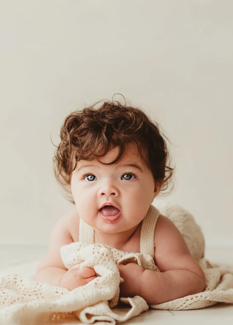 A baby with curly brown hair and blue eyes lying on a cream-colored blanket, looking surprised or curious.