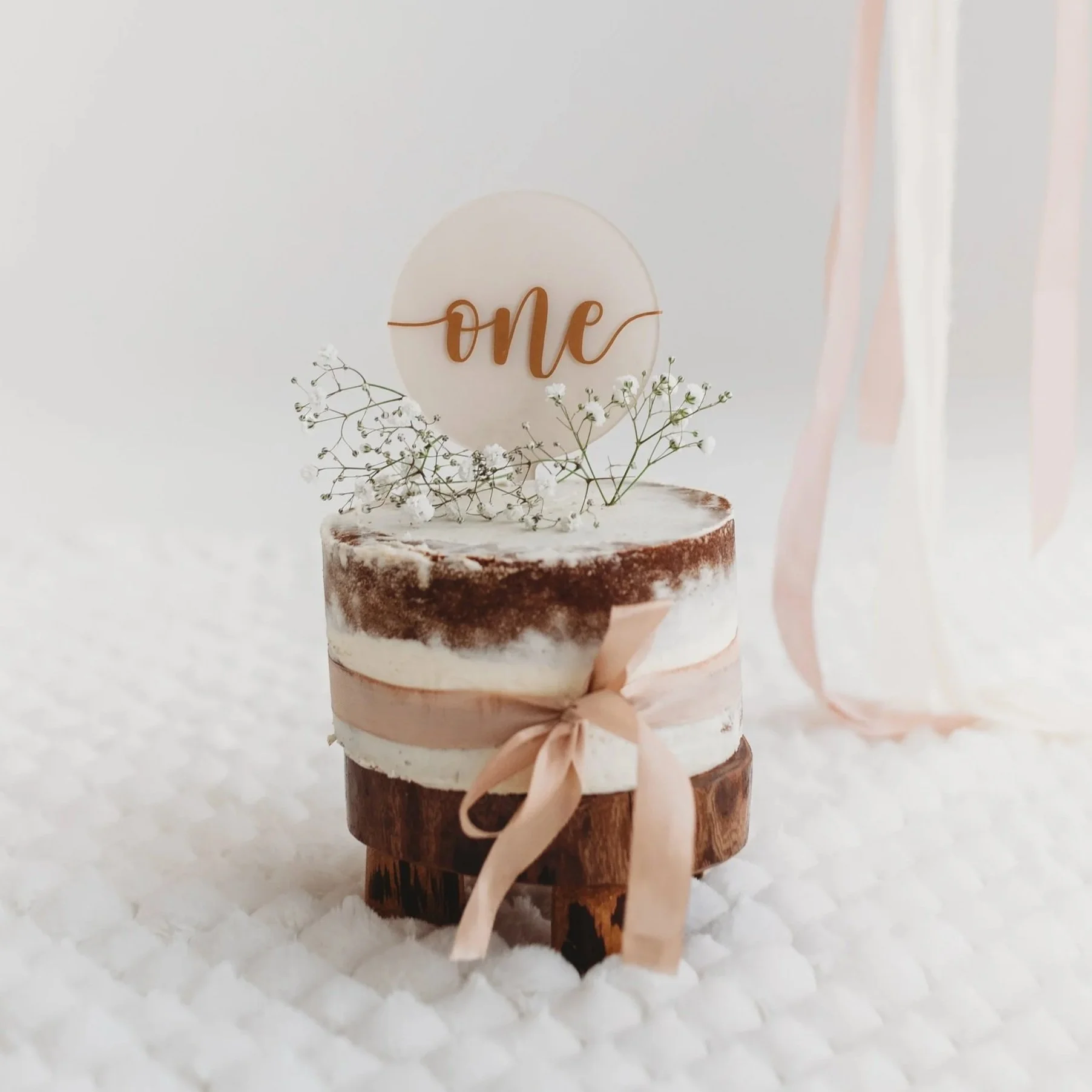 Wooden log with white frosted cake on top, decorated with small white flowers and a tan ribbon. A white circular cake topper with the word 'one' written in tan cursive lettering.