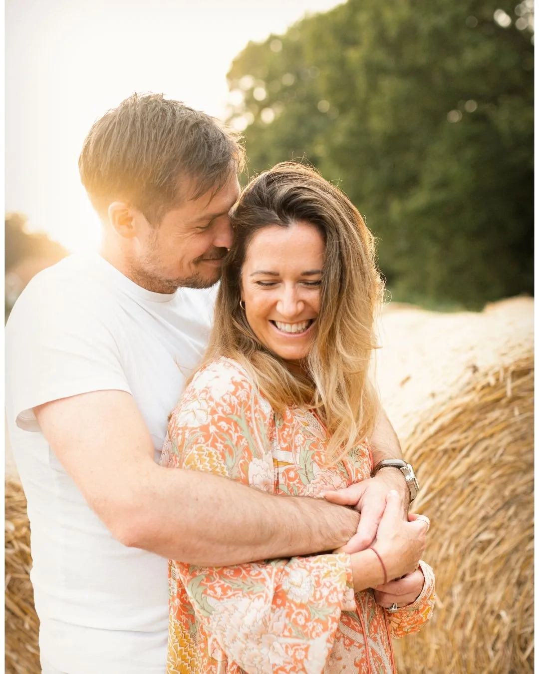 A smiling woman with long, wavy hair embracing a man from behind in a field with hay bales, during golden hour.