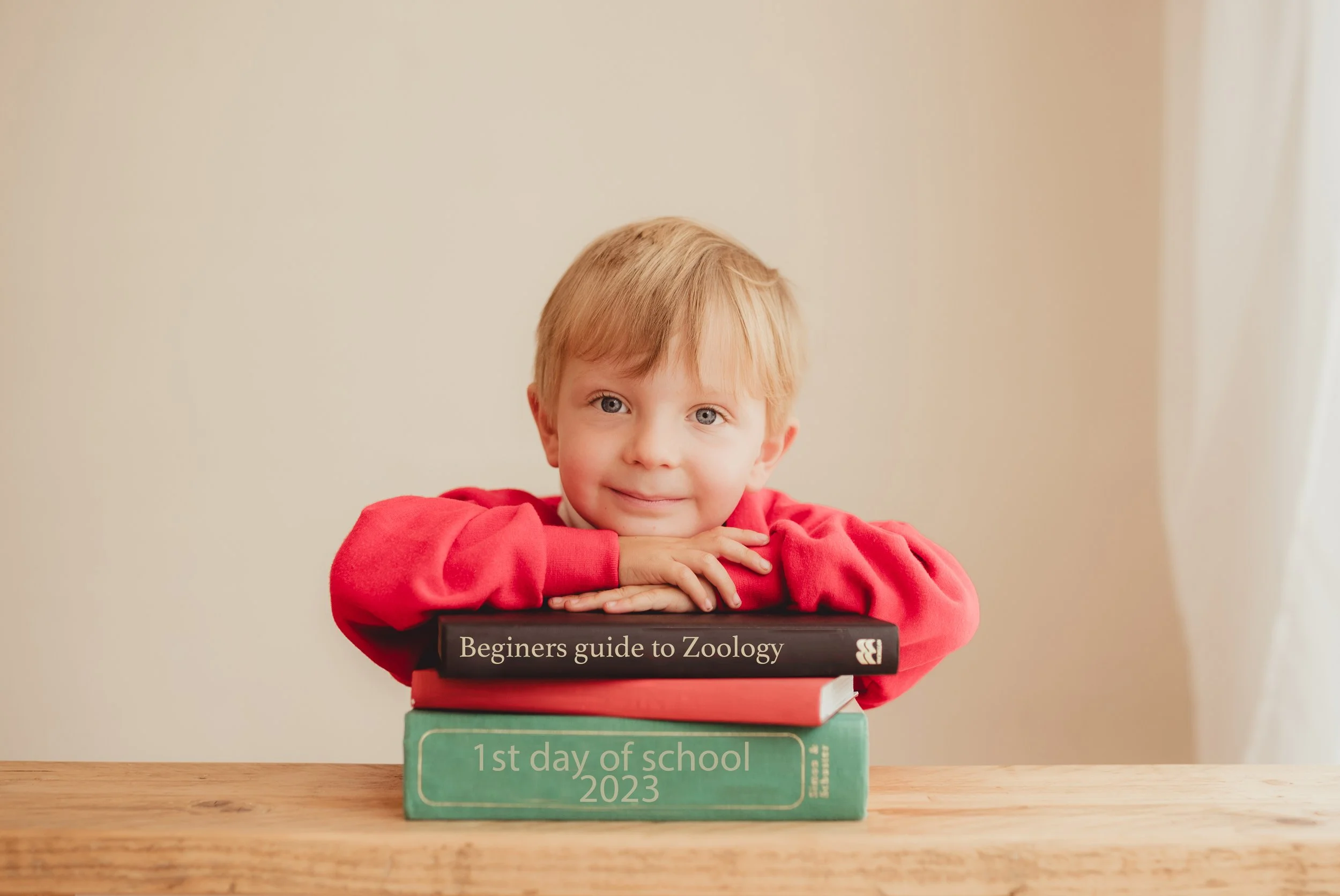 A young boy with blond hair and blue eyes resting his chin on folded arms on top of a stack of three books. The top book is titled 'Beginners guide to Zoology', and the bottom book is titled '1st day of school 2023'. The boy is wearing a red long-sle