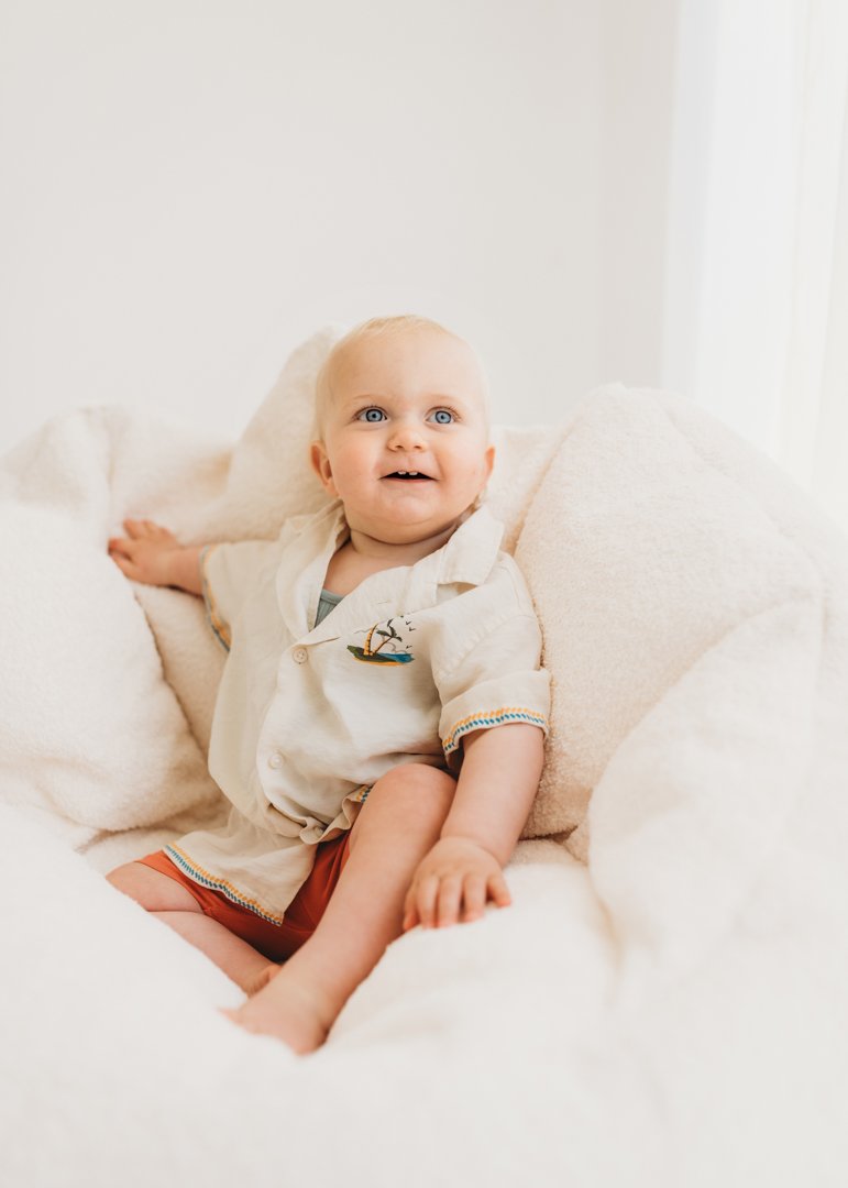A young child with blonde hair and blue eyes sitting on a white bed surrounded by white pillows, wearing a cream-colored shirt with embroidery and orange shorts, smiling and looking at the camera.