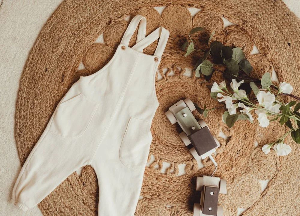 Cream-colored baby overalls on a woven circular rug with electrical cords and a branch of white flowers and green leaves.