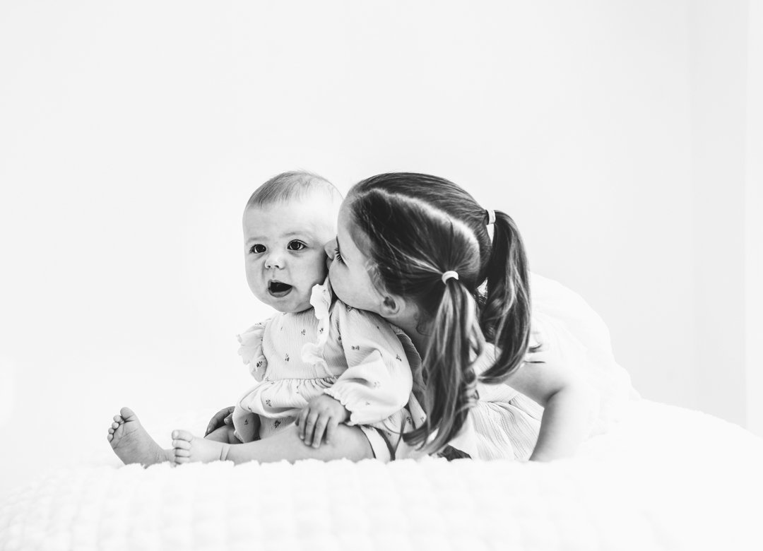 A young girl is kissing a toddler on the cheek while lying on a bed in a bright room.