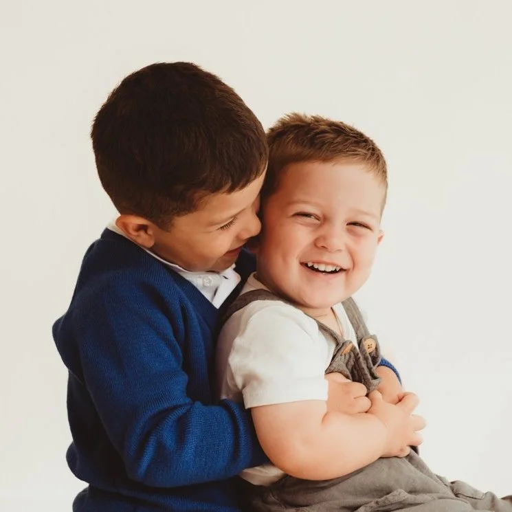 Two young boys laughing and hugging each other, one wearing a blue sweater and the other in a white shirt and gray overalls.