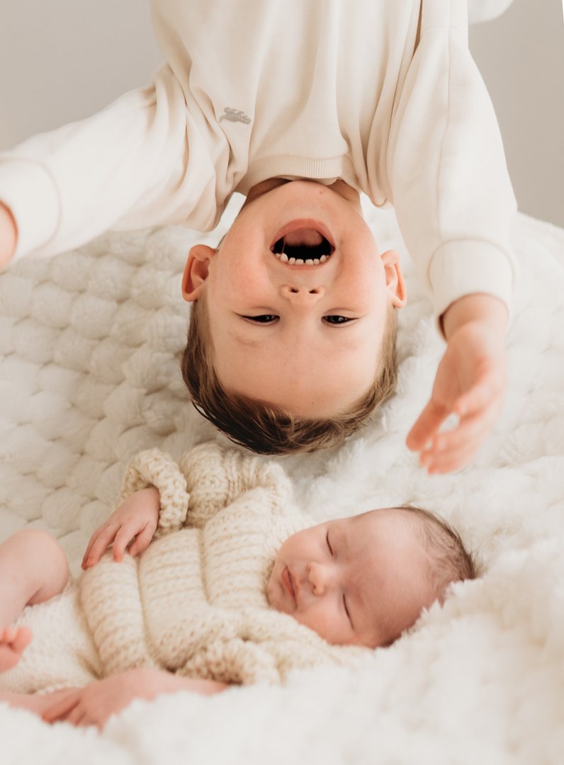 Toddler laughing upside down over the top of his newborn sister as she sleeps during her newborn photoshoot Saffron Walden Essex