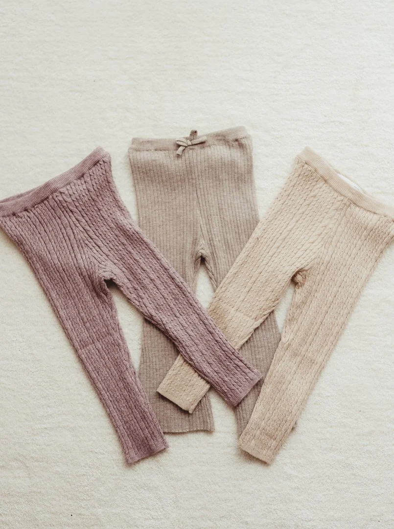 Three pairs of knit leggings in soft pink, beige, and cream colors arranged on a light surface.