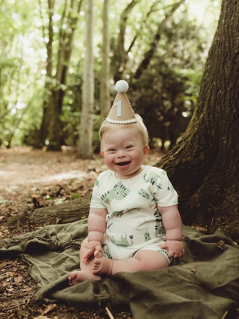 A cute and smiling one year old with a birthday hat on in the woodland for a cake smash photoshoot
