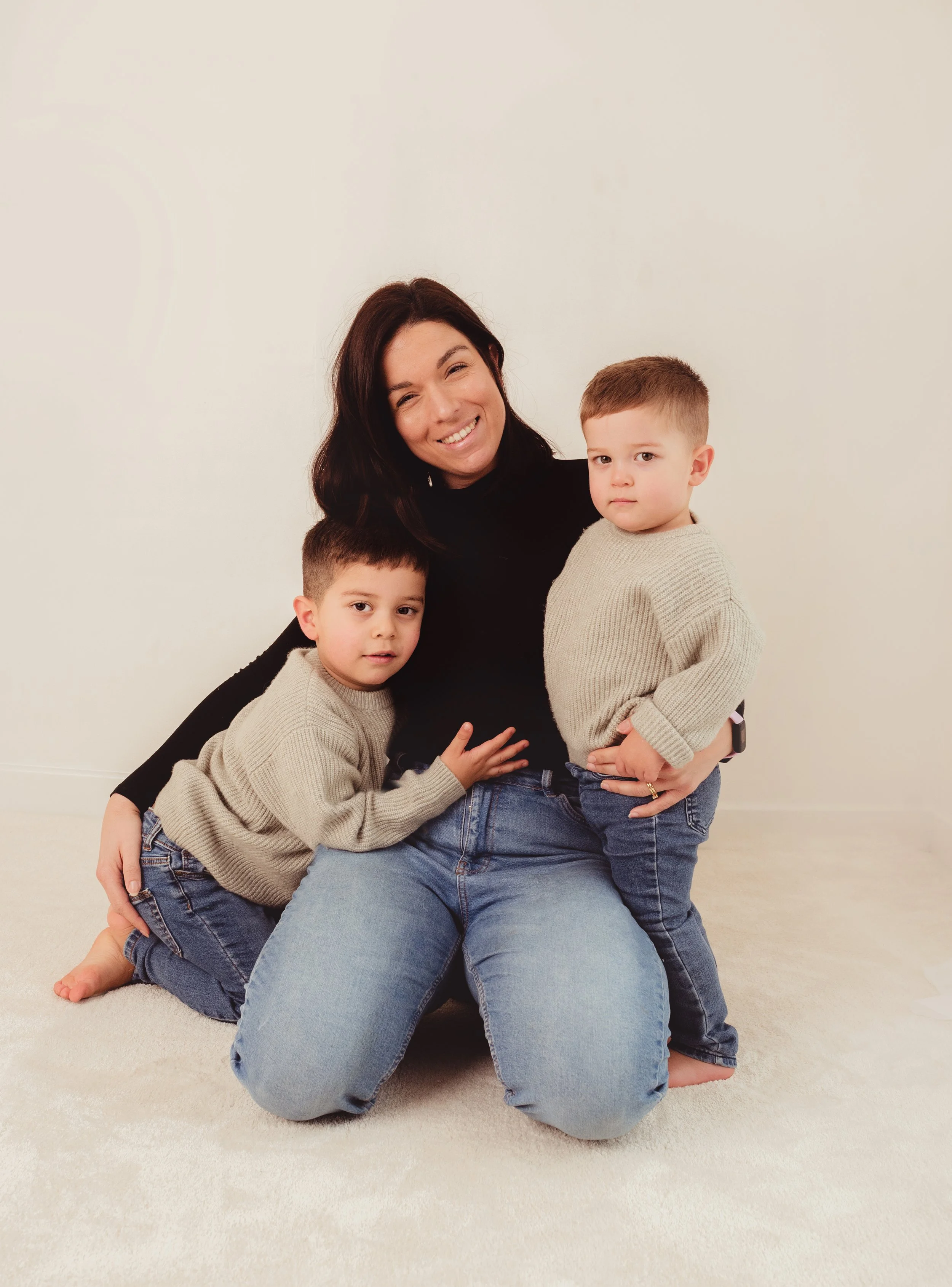 A woman smiling with two young boys on a beige carpeted floor against a plain white wall.