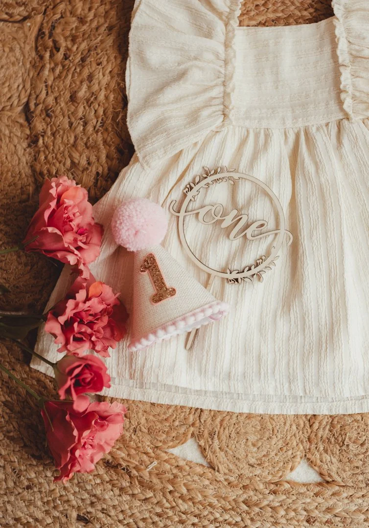 A white dress, pink roses, a white cone-shaped birthday hat with a rose gold number 1, a pink pom-pom, and a wooden 'love' sign on a woven surface.
