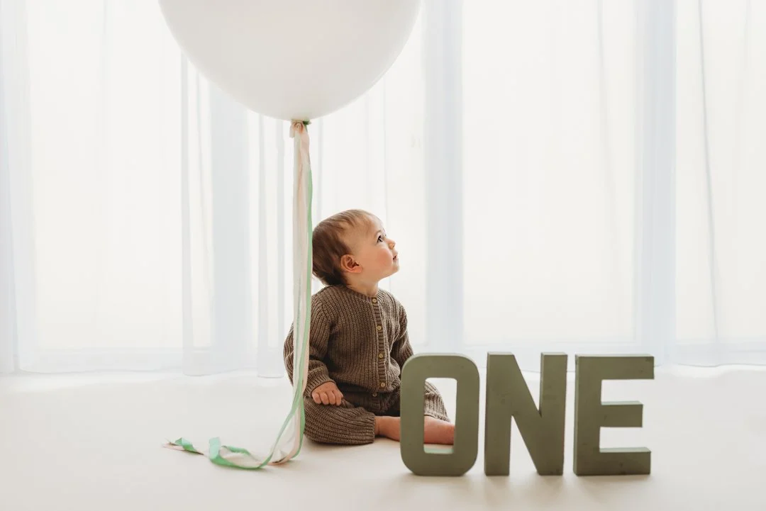 A young child sitting on a white surface near a white balloon and large block letters spelling 'ONE', with a bright background and sheer curtains.