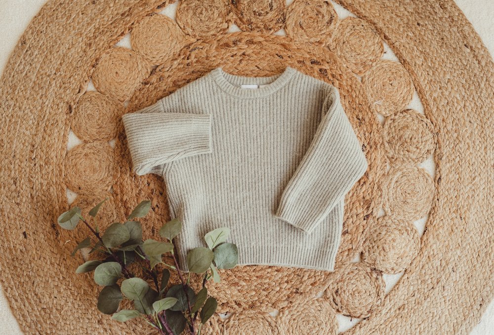 A beige, chunky knit sweater laid flat on a circular woven jute rug. A sprig of eucalyptus leaves is placed beside the sweater.