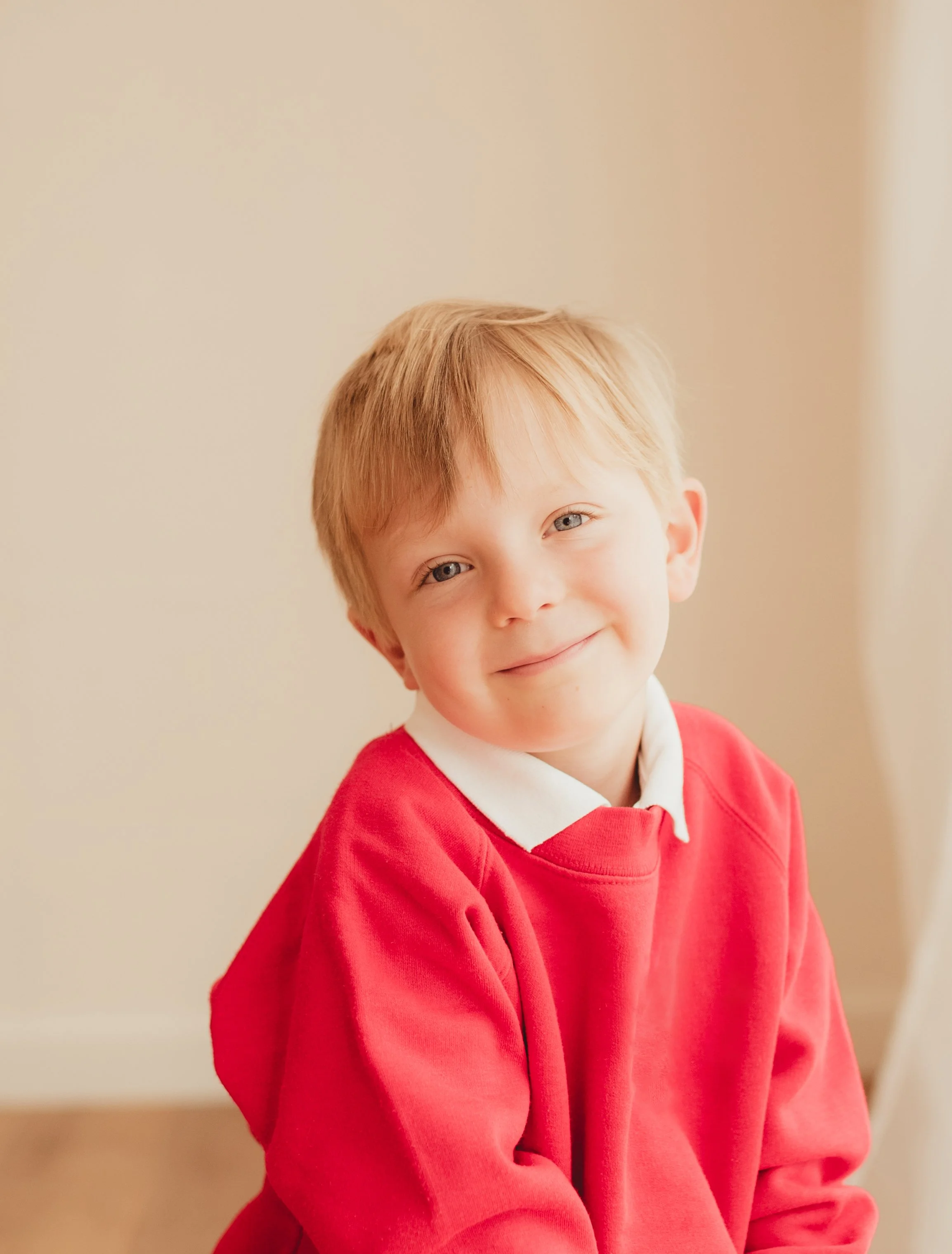 A young boy with light red hair, blue eyes, and a light complexion, smiling softly, wearing a red sweater over a white collared shirt, standing against a neutral beige background.
