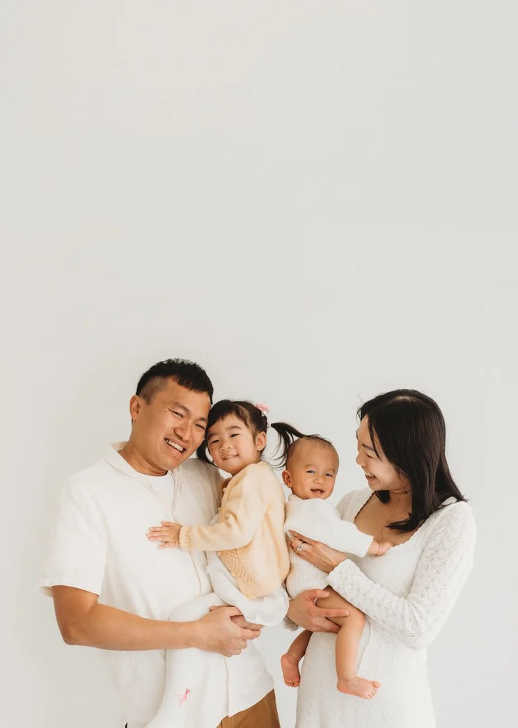 Happy family of four, parents and two young children, smiling and looking at each other against a plain white background.