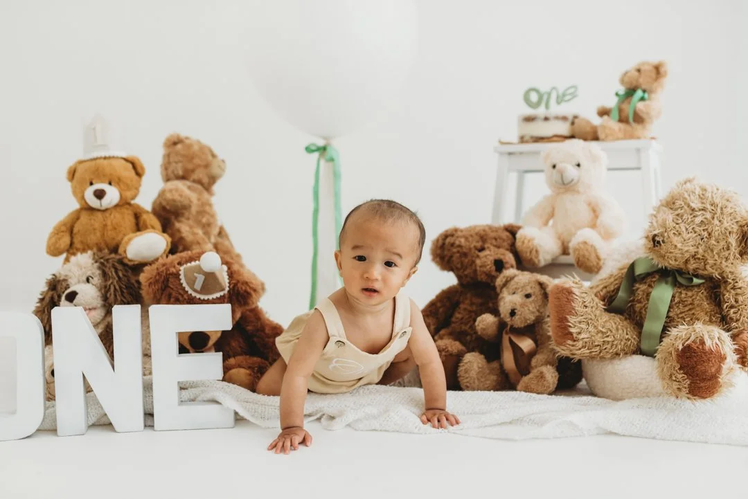 A baby in a beige outfit crawling on a white blanket surrounded by teddy bears and decorative items for a first birthday celebration with Jodie Wilson Photography in Saffron Walden Essex