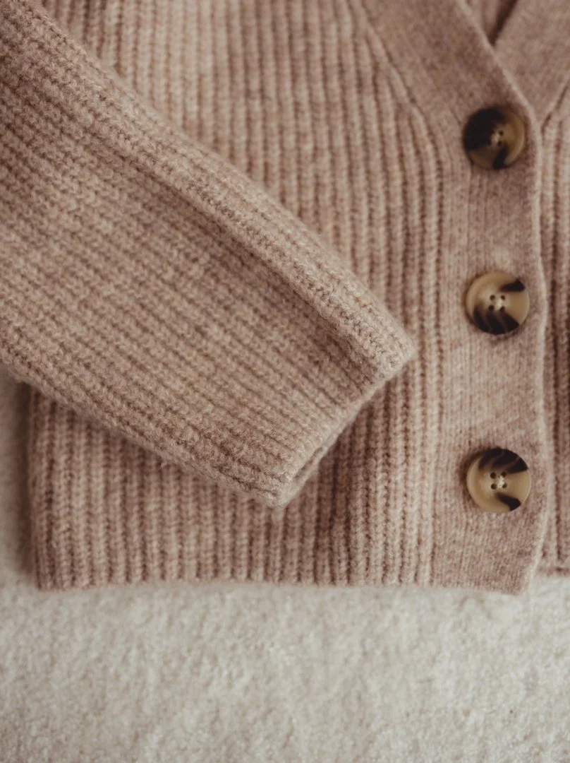 Close-up of a beige, ribbed knit cardigan with tortoiseshell buttons resting on a soft, cream-colored surface.