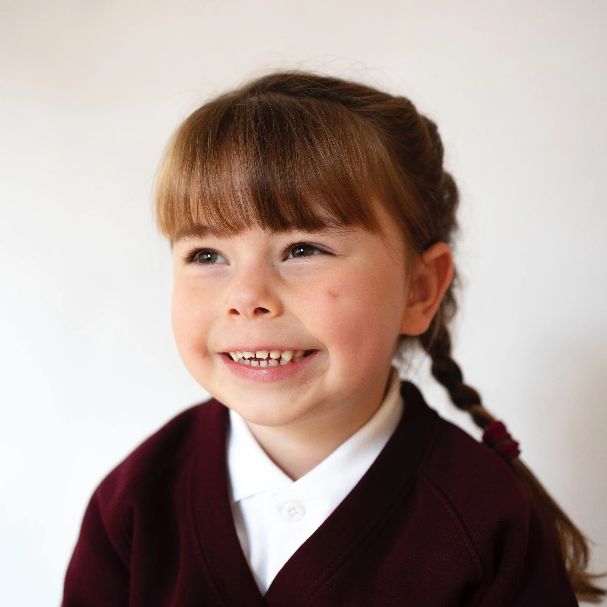 Close-up of a young girl with brown hair, wearing a white shirt and dark maroon sweater, smiling and looking upward.