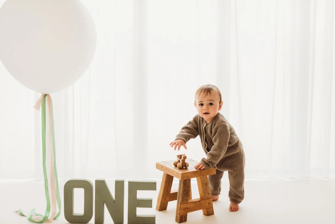 A baby standing on the floor next to a small wooden table, a large white balloon with colorful ribbons, and the letters 'ONE' in front of the table, celebrating their first birthday.