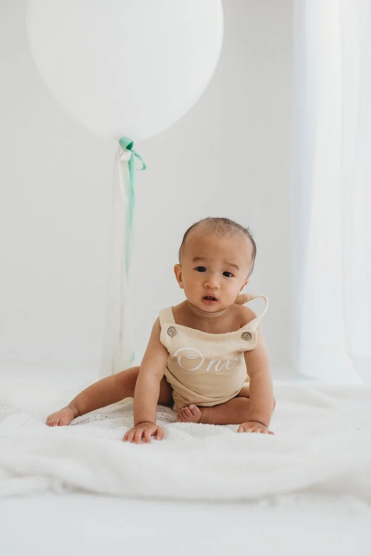 A baby sitting on a white blanket in front of a white wall with a large white balloon tied with a ribbon in the background.
