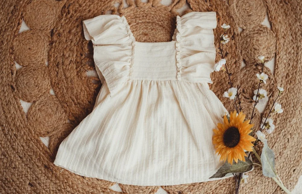 A cream-colored, ruffled sundress with puff sleeves on a textured, knobby, brown surface, next to a sunflower and some small white flowers.