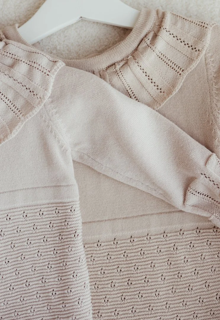 Close-up of a beige sweater with a textured, knitted pattern on the collar and lower part, on a white hanger against a light background.