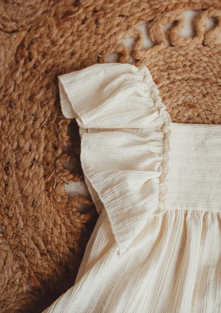 Close-up of a cream-colored dress with puffed ruffled sleeves, placed on a woven straw surface.