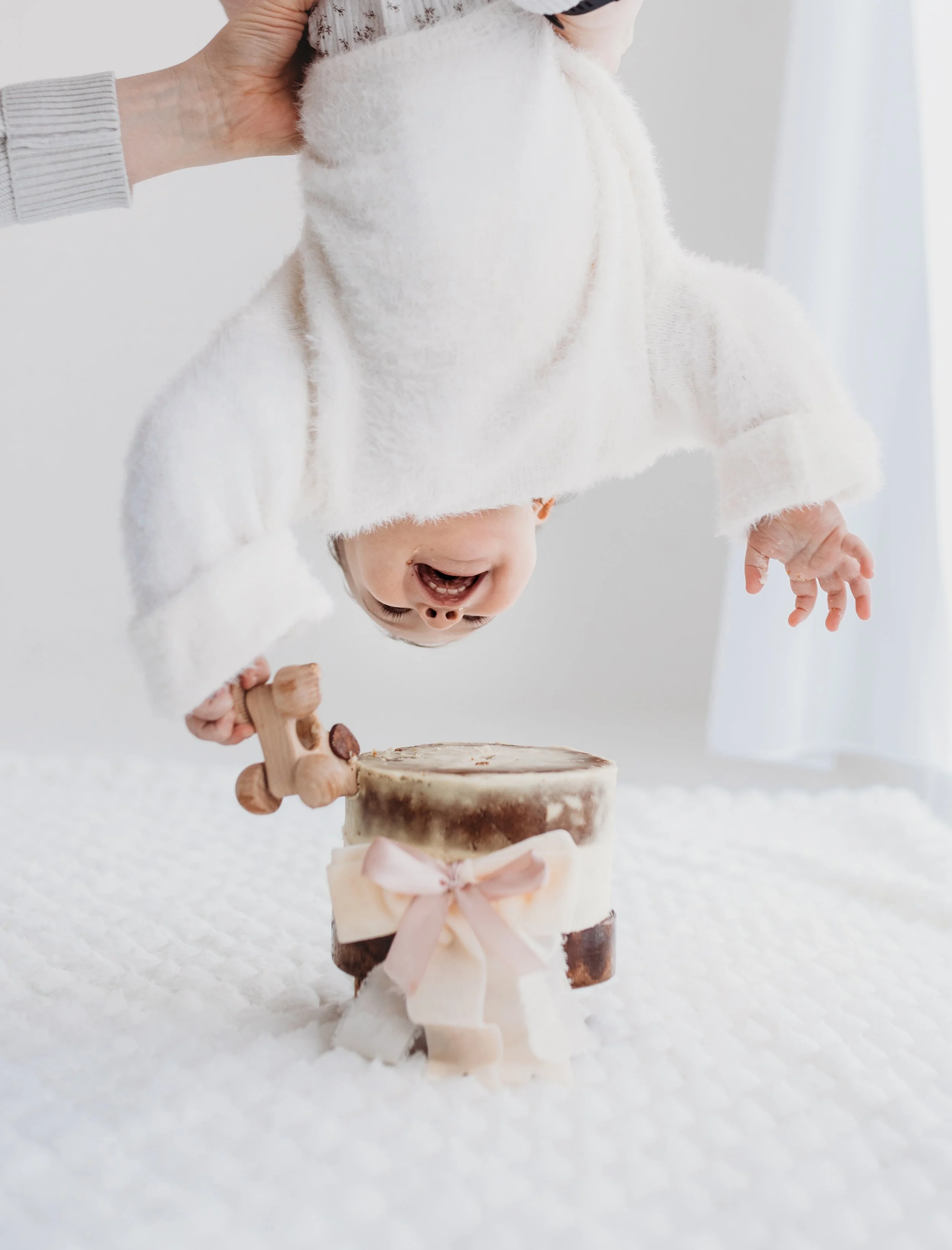 faceless image of a Child in a white jumper playing at a cake smash, holding a small wooden dog and standing in front of a layered cake decorated with a pink ribbon.