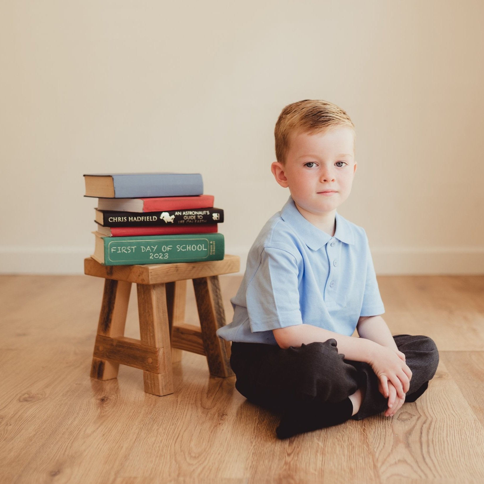 A young boy with red hair sitting cross-legged on a wooden floor, next to a small wooden stool with a stack of books, including one titled 'First Day of School 2023'. The boy is wearing a light blue collared shirt and dark pants, looking slightly to 