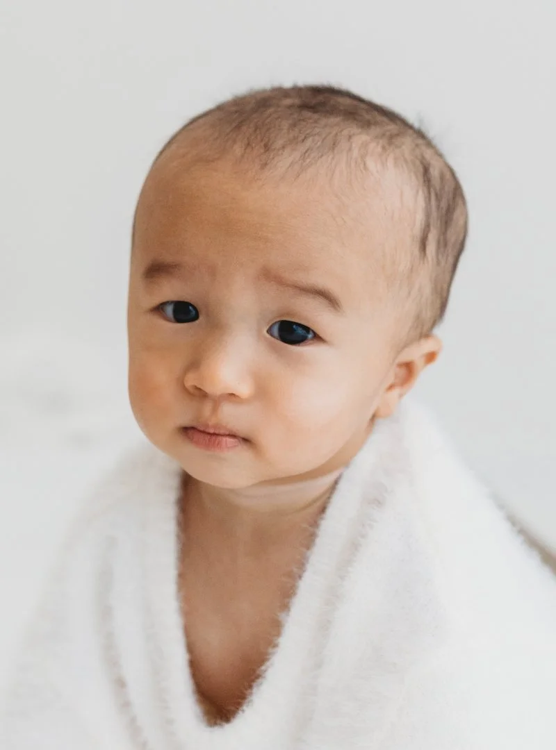 A young Asian boy with a shaved head looking slightly to the side, wrapped in a white towel.