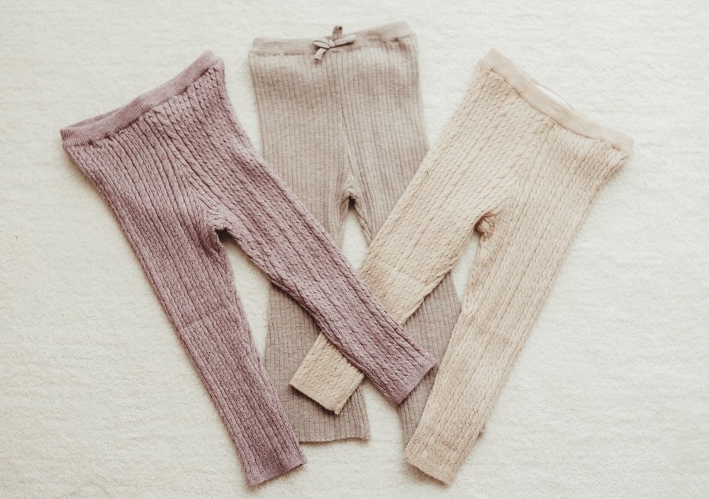 Three pairs of ribbed knit tights in purple, beige, and cream colors are laid flat on a light background.