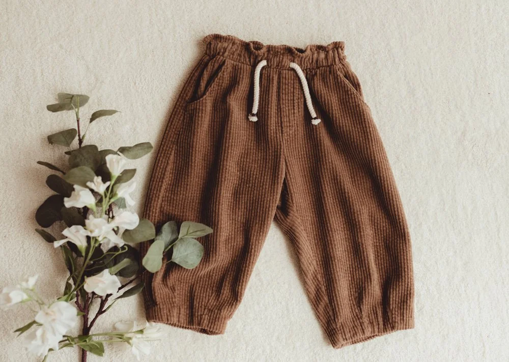 Brown corduroy pants with an elastic waistband, white drawstring, and patch pockets, laid on a light-colored surface next to a bunch of white flowers.