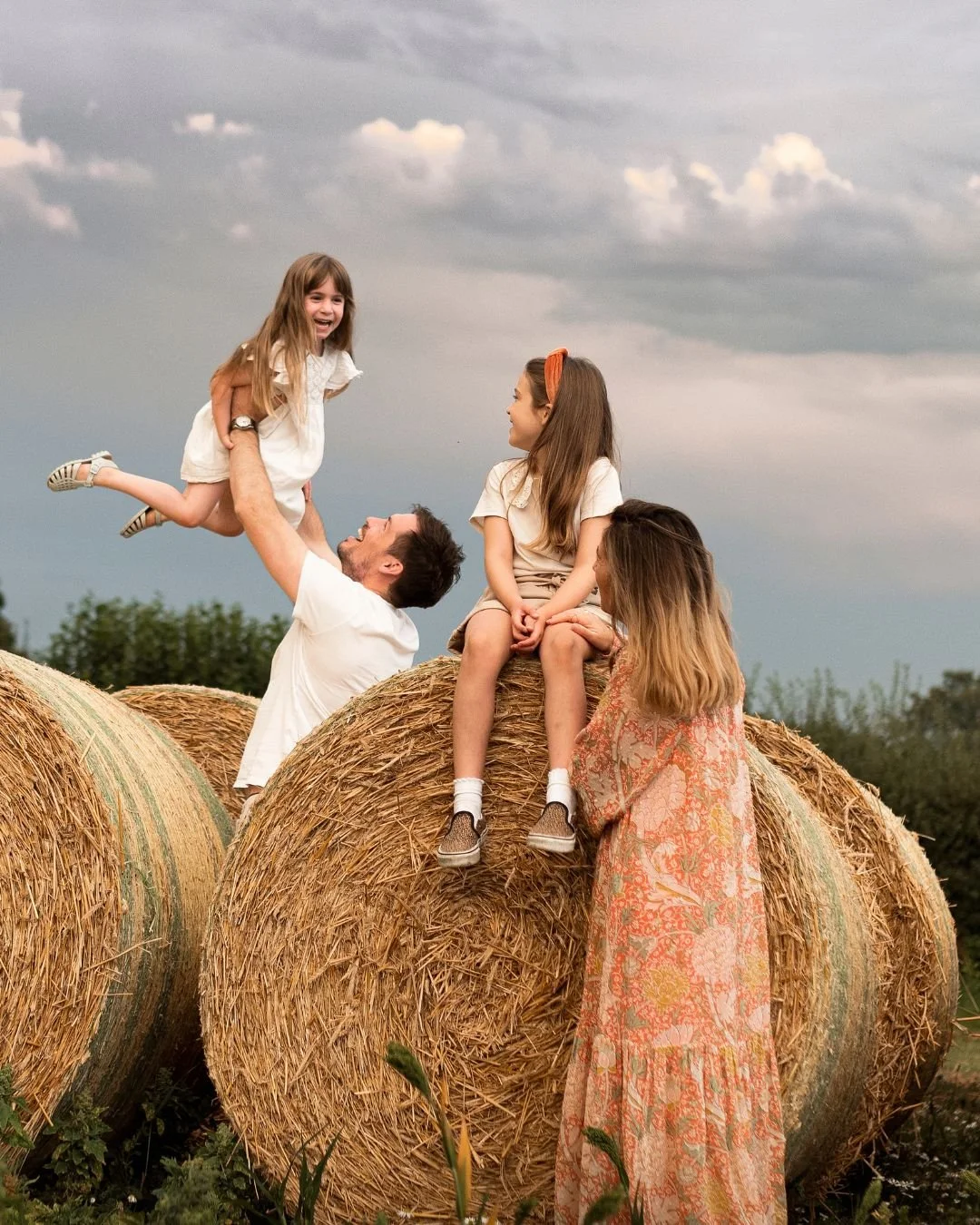Two young girls sitting on top of a hay bale and a man lifting a girl in the air, all smiling and enjoying outdoors on a cloudy day.