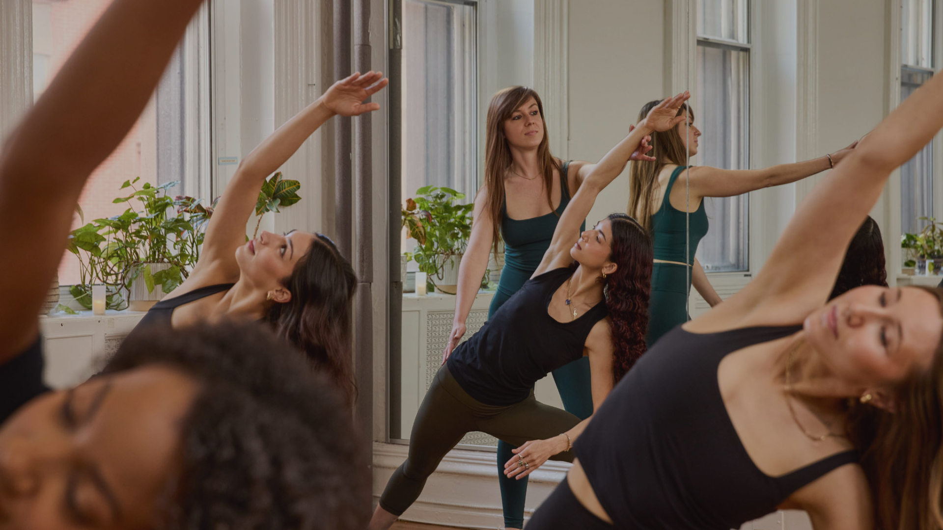 Women participating in a yoga class in a bright studio with large windows, plants, and mirrors, all wearing athletic clothing and stretching.
