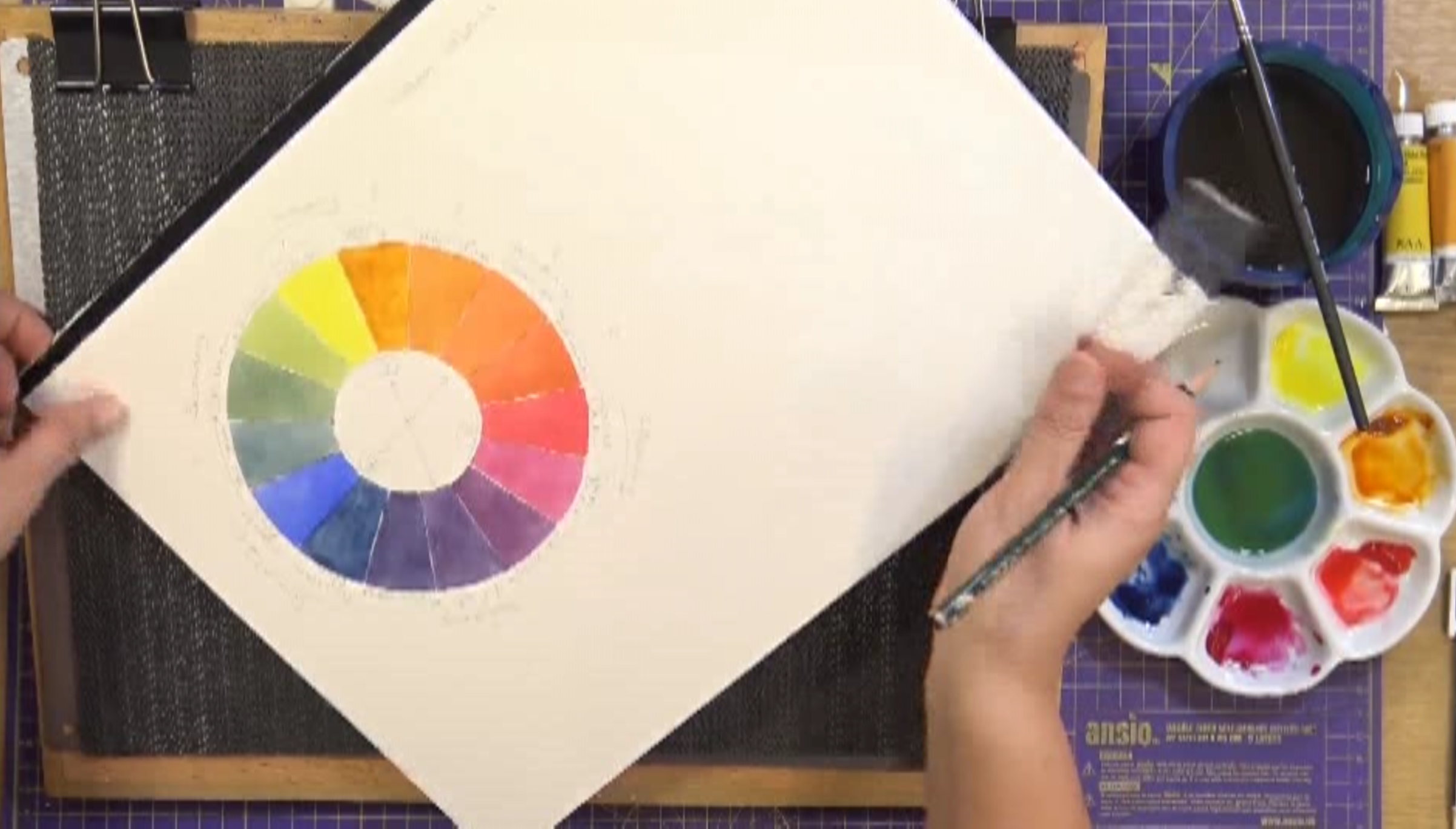 A person holding a color wheel on a sketchpad, next to a paint palette with various colors, brushes, and art supplies on a crafting mat.