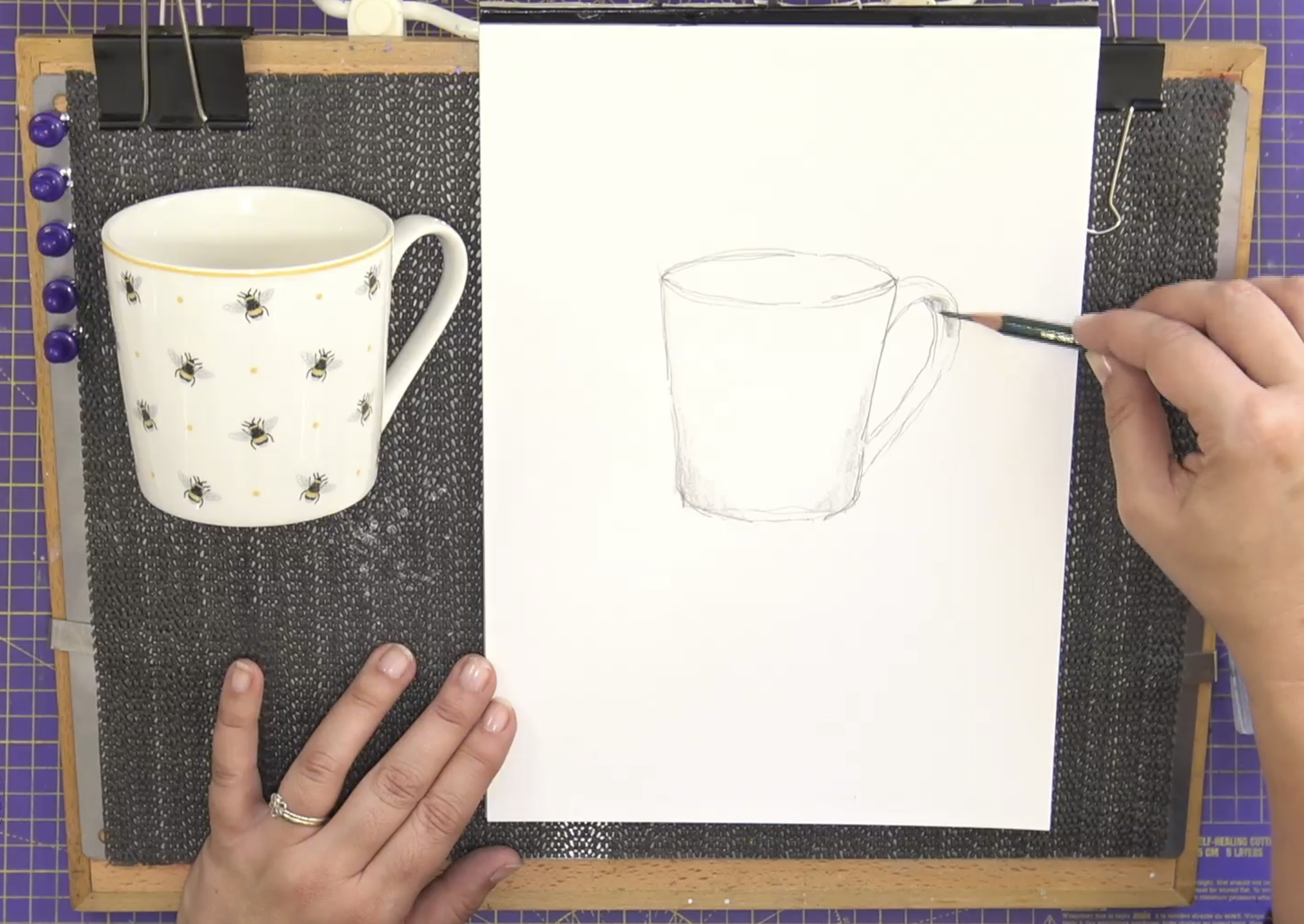 Person sketching a mug on white paper next to a real mug with bee pattern on a textured surface.