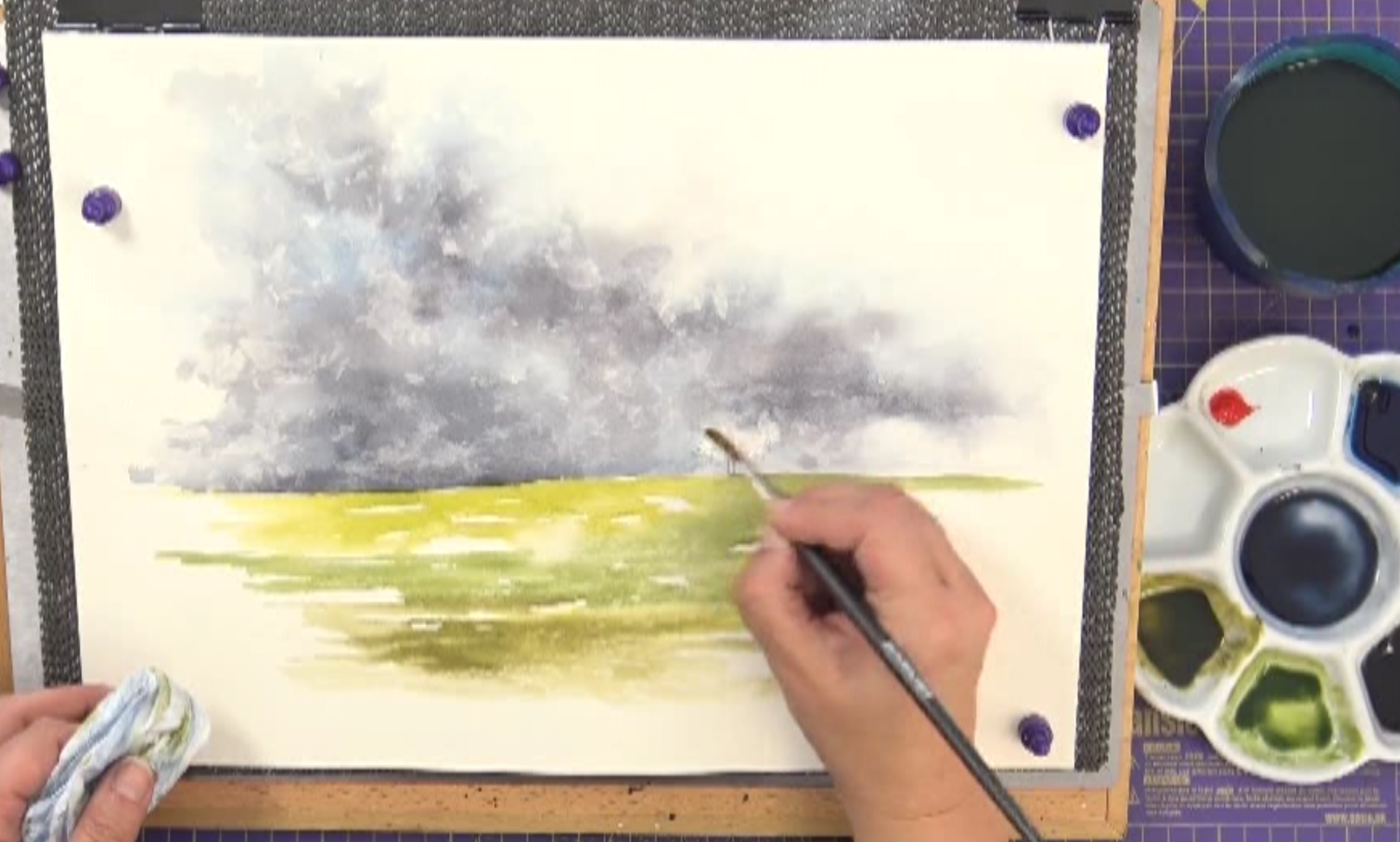 Person painting a watercolor landscape with dark stormy clouds and grassy field, using a paintbrush and palette.