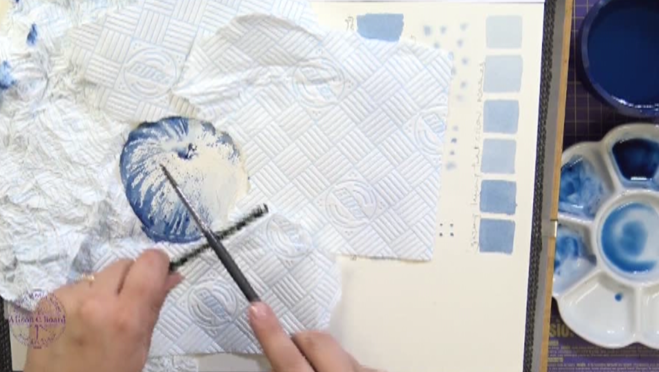 Person painting with watercolors on a textured paper using blue shades, surrounded by a color palette and paper towels.