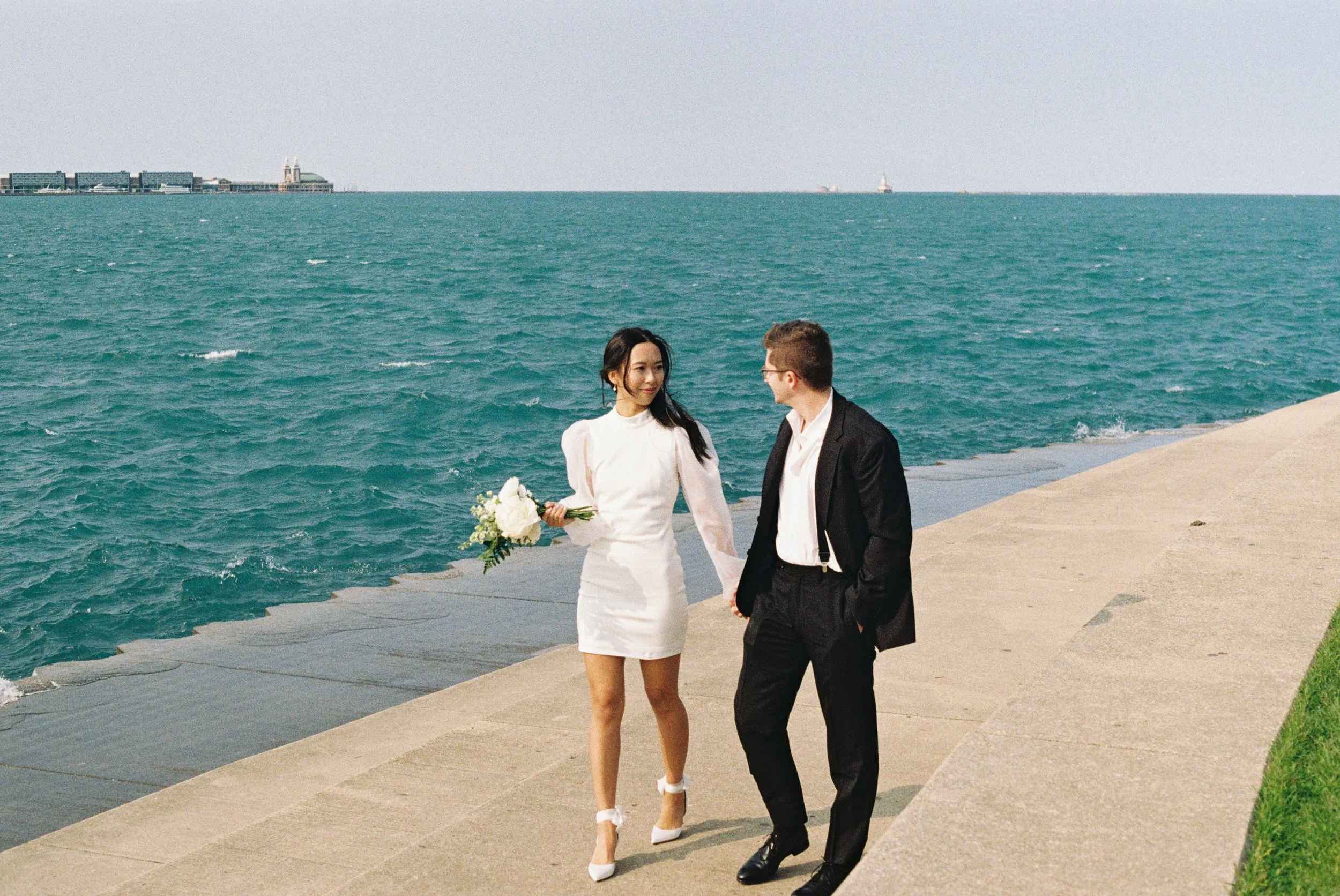 A woman in a white dress holding a bouquet walks with a man in a black suit along a waterfront promenade, with the ocean and a city skyline in the background.