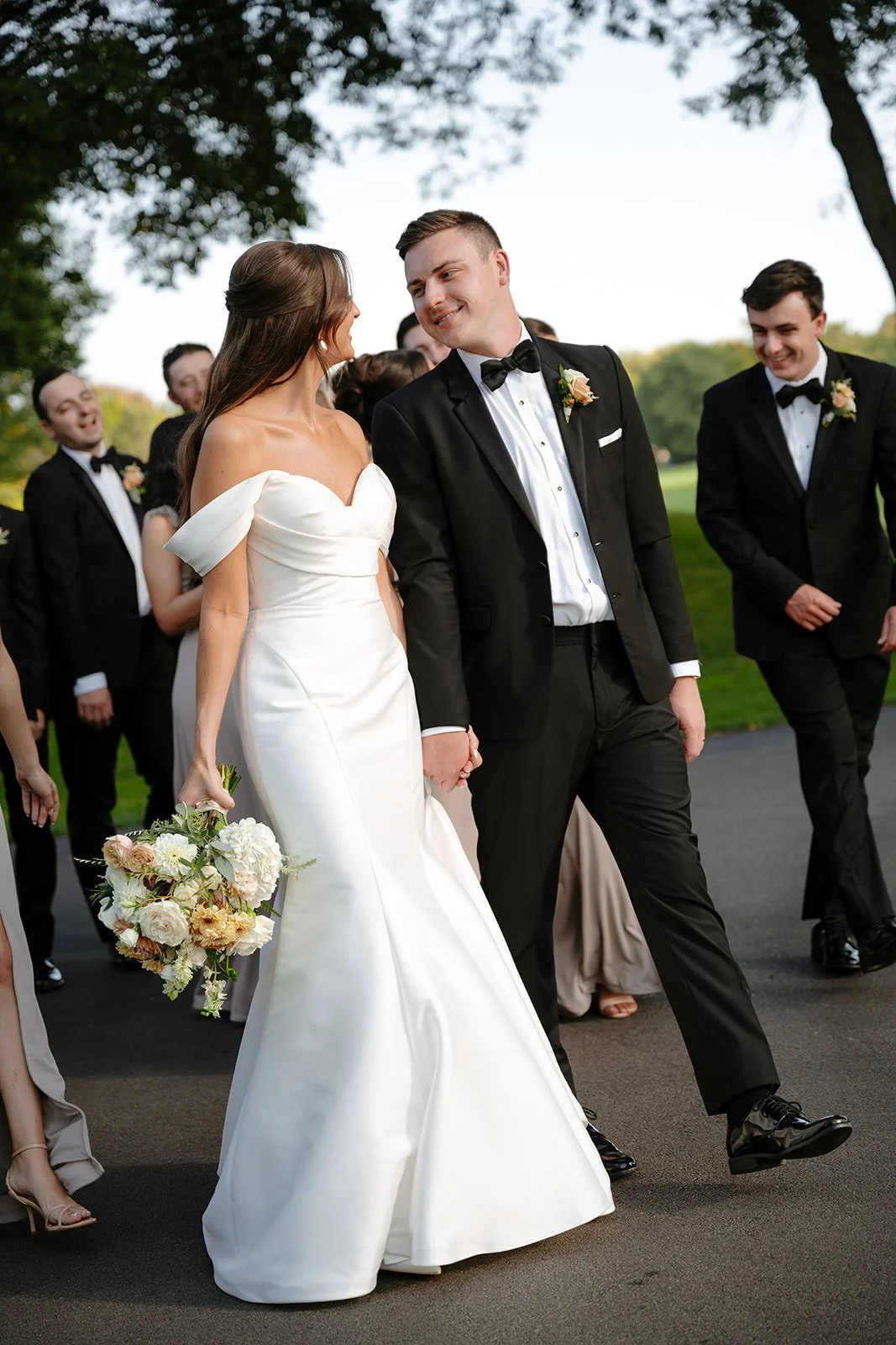A bride and groom holding hands, walking outdoors with wedding guests in the background, on a paved path near trees.