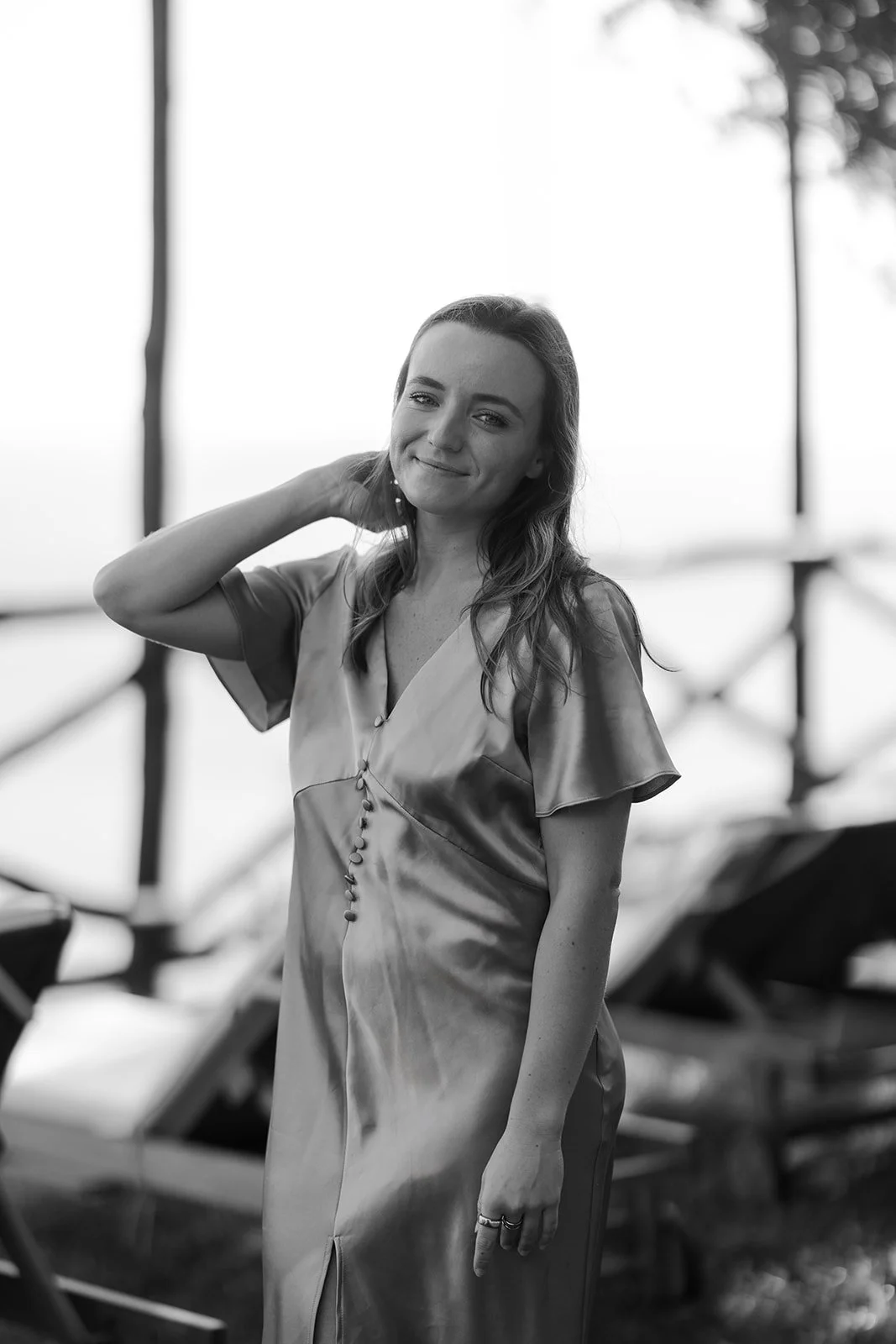 A woman in a silky dress standing outdoors near a beach, with a relaxed expression and one hand touching her neck, in a black and white photograph.