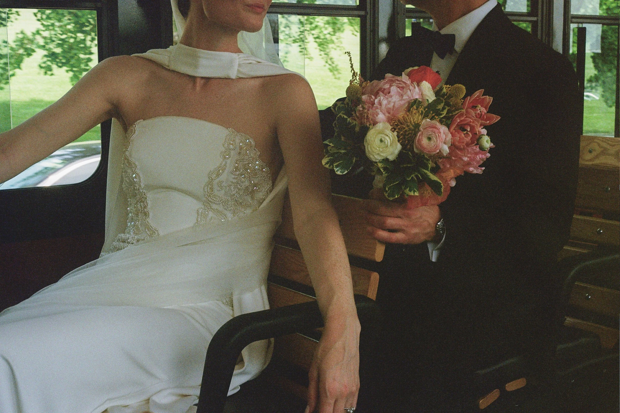 A bride in a white wedding gown sitting on a bench with a groom in a black tuxedo holding a bouquet of pink and white flowers.