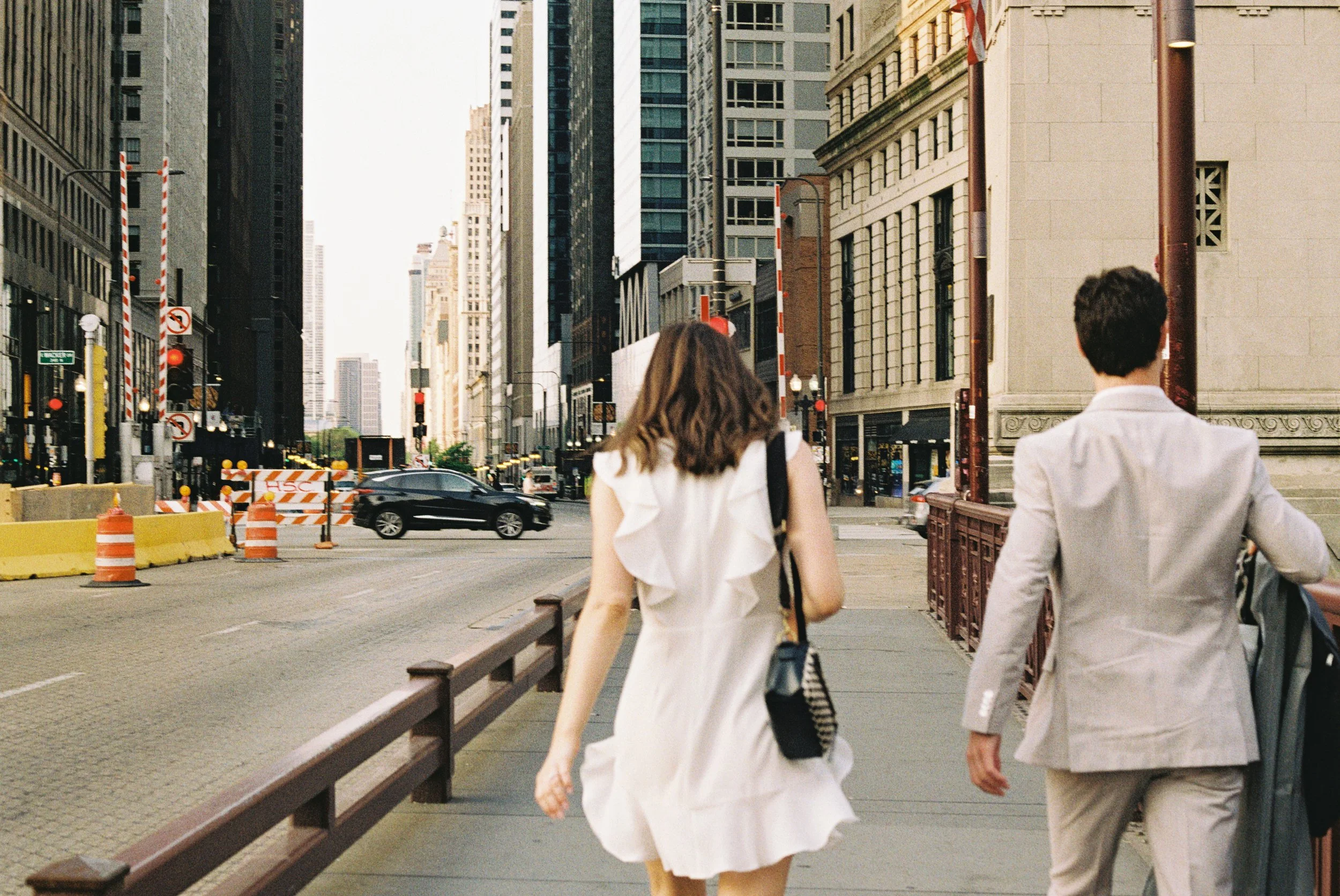 Two people walk on a city sidewalk with tall buildings, traffic lights, and cars in the background.