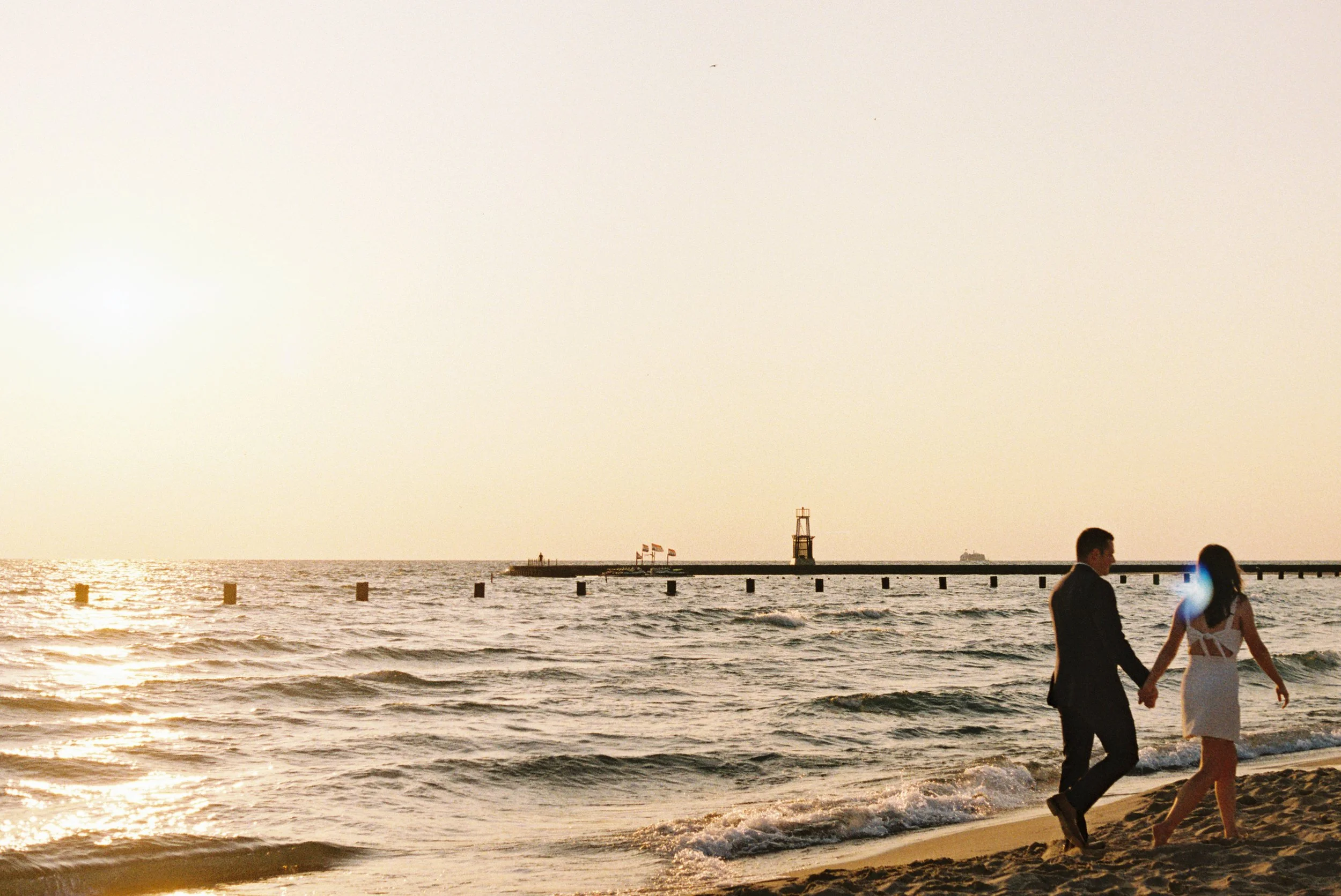 A couple in formal attire walking hand in hand on a beach at sunset, with the ocean, a breakwater, a lighthouse, and flags in the background.