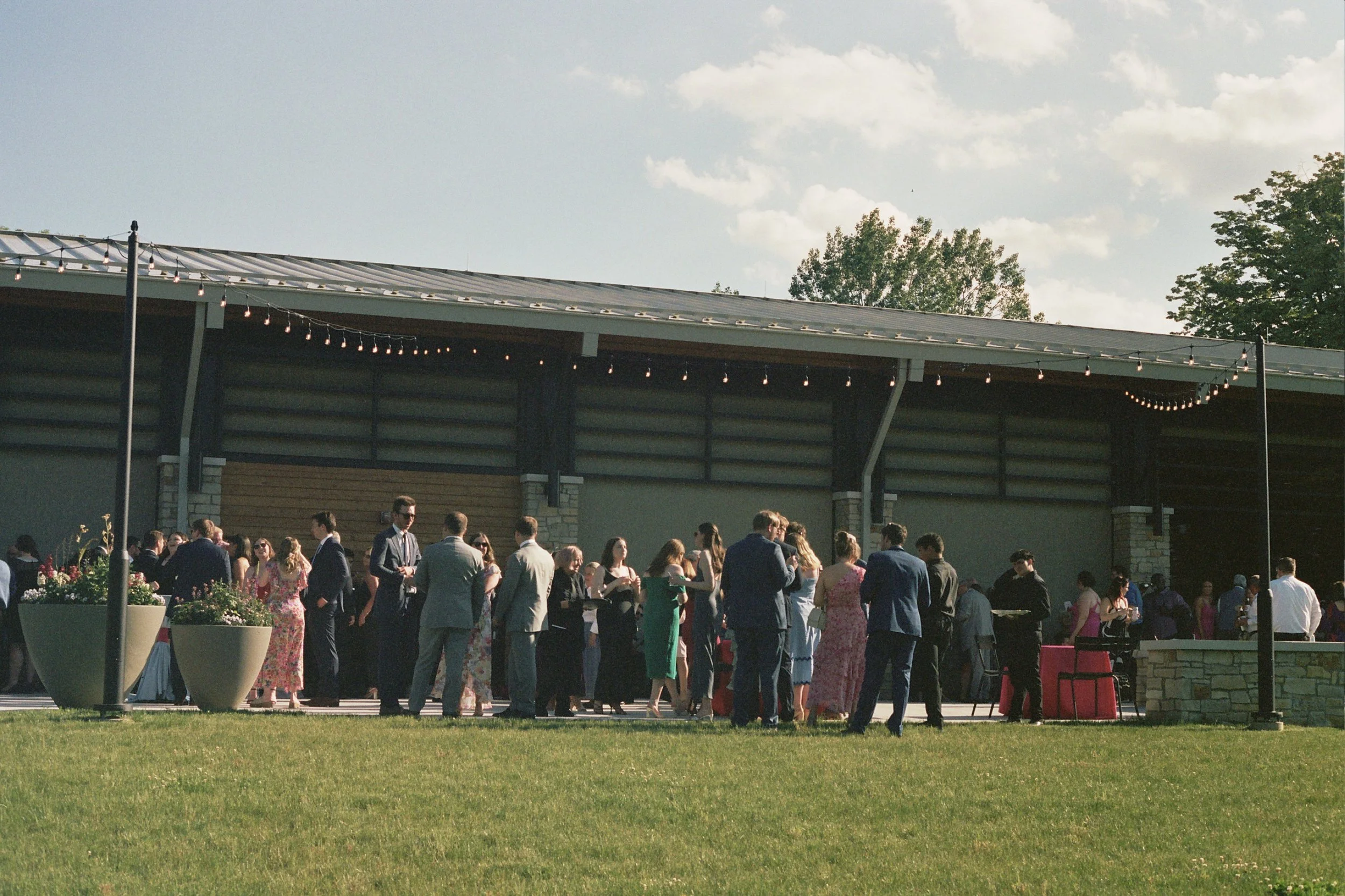 People gathered outdoors at a social event, with some standing and talking, under string lights near a building with a metal roof and trees in the background.