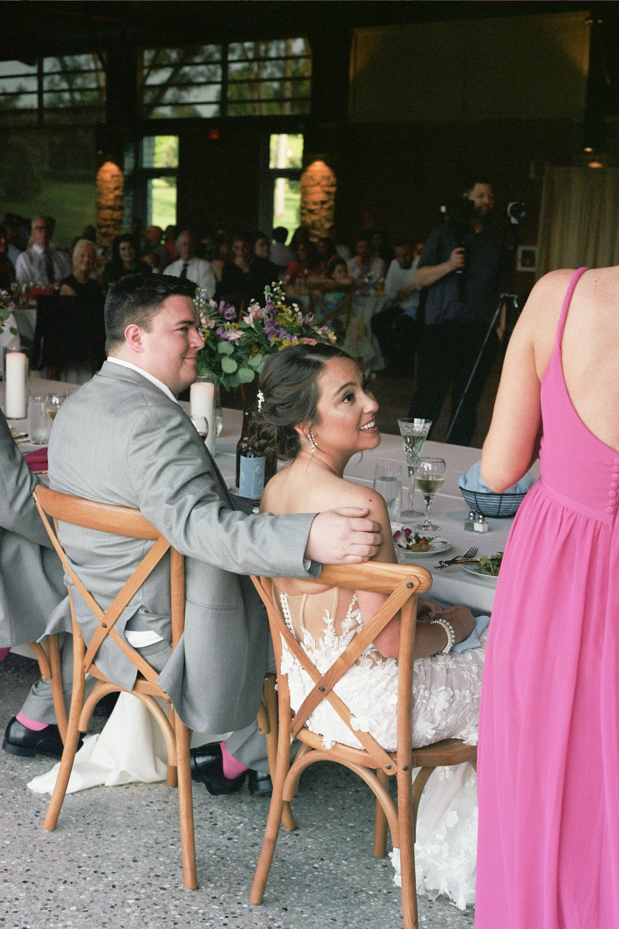A bride and groom sitting at a wedding reception table, with the bride smiling and looking at a woman in a pink dress standing nearby. The reception is indoors with many guests in the background.