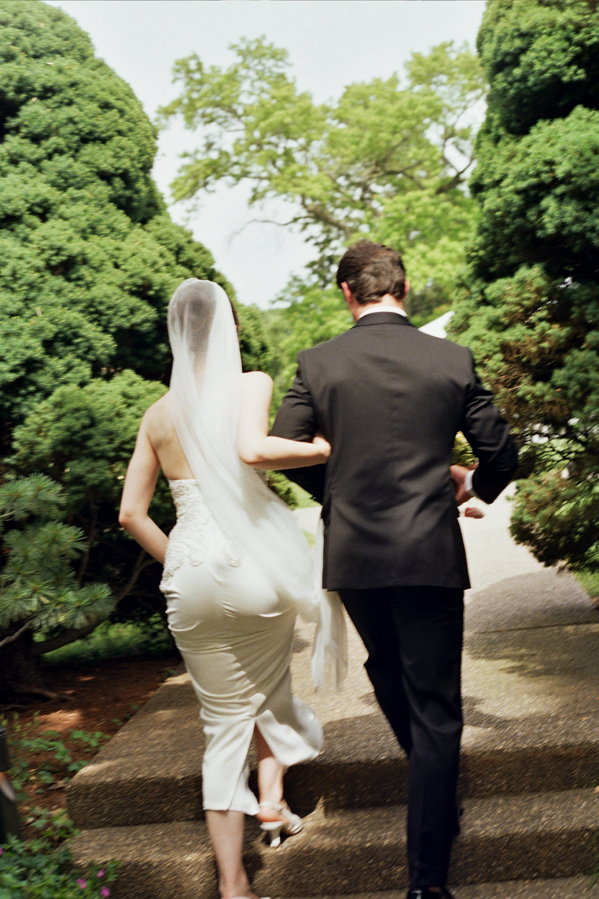A bride in a white wedding dress and veil walking arm in arm with a groom in a black suit on a stone path surrounded by green trees.