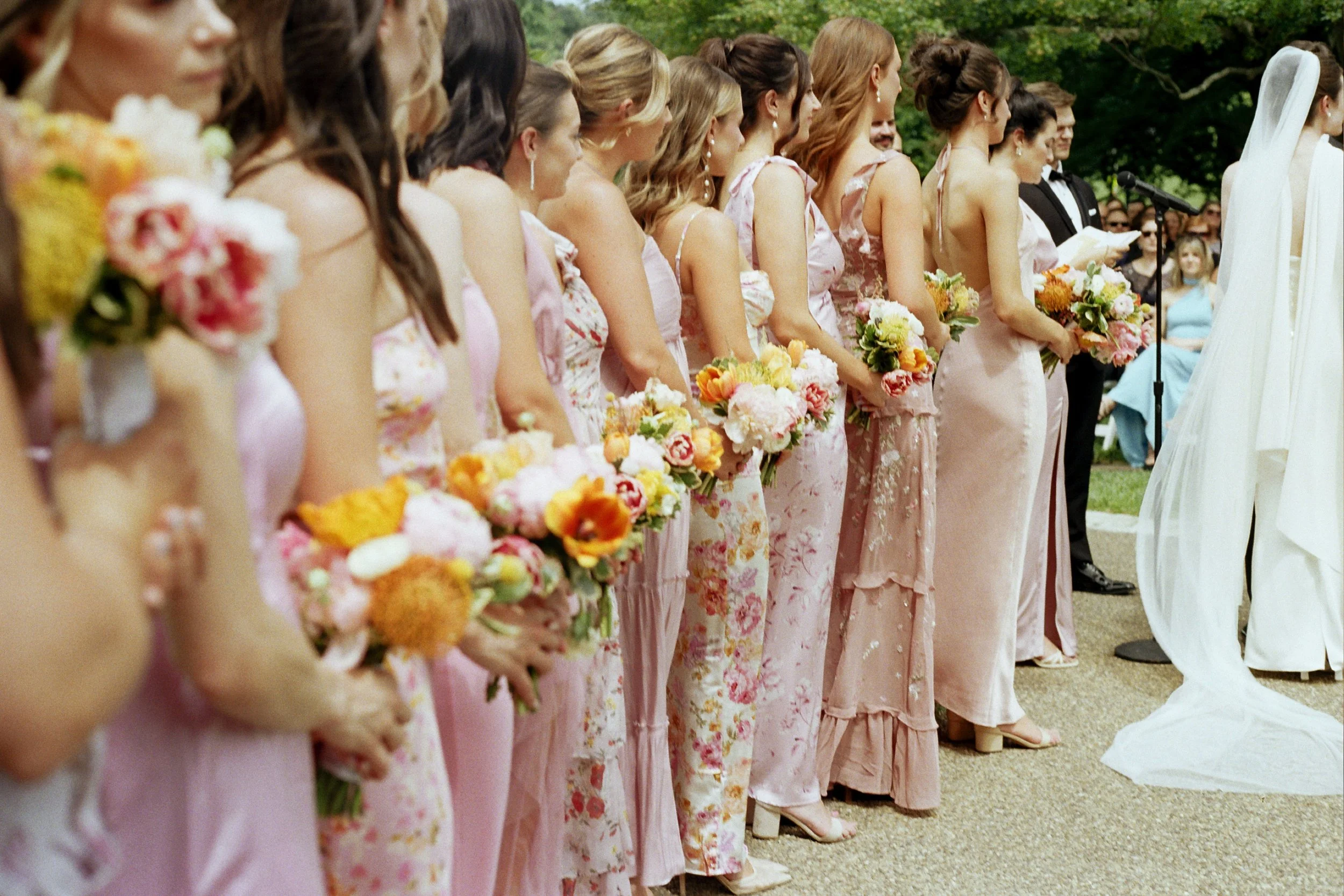 Line of bridesmaids in pink floral dresses holding bouquets at a wedding ceremony outdoors.