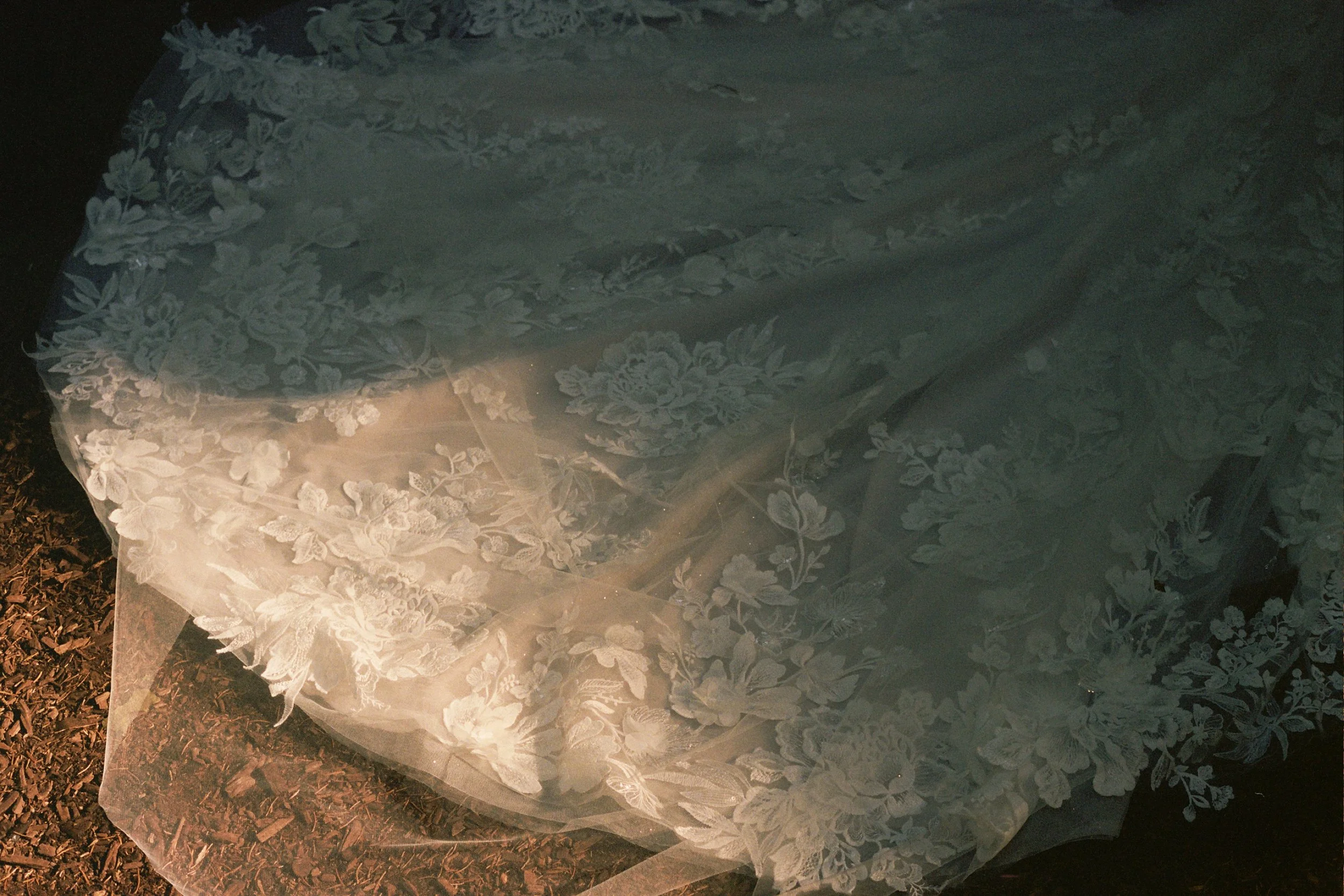 A table covered with a lace tablecloth with floral embroidery, partially illuminated, with a rough soil ground visible at the bottom left.
